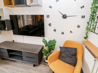 A living area with a wall-mounted TV above a low cabinet a large clock on the wall an orange chair with a black cushion and green plants at 20 Bishops Way in Falmouth