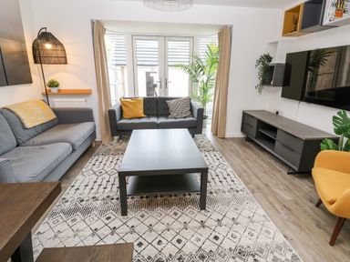 A living room with two grey sofas a yellow armchair a coffee table a patterned rug a TV on a stand and large windows at 20 Bishops Way Falmouth