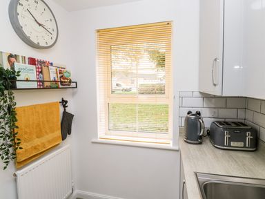 A kitchen with a window with blinds a kettle toaster wall clock and books on a shelf at 20 Bishops Way in Falmouth
