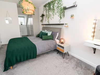A bedroom with a bed featuring green and grey bedding a small nightstand with a lamp hanging plants and a vanity mirror at 20 Bishops Way in Falmouth