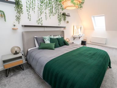 A bedroom with a grey bed and green pillows and blanket with hanging plants and a lit vanity mirror at 20 Bishops Way in Falmouth