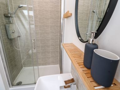 A bathroom with a shower enclosure a white sink a round mirror and a wooden shelf with soap dispenser and cup at 20 Bishops Way in Falmouth