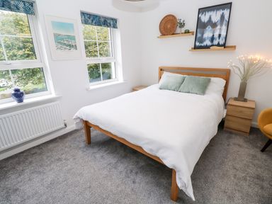 A bedroom with a wooden bed and white bedding two green pillows two windows with blue curtains a wooden nightstand with a lamp a yellow chair and wall shelves with decorative items at 20 Bishops Way in Falmouth
