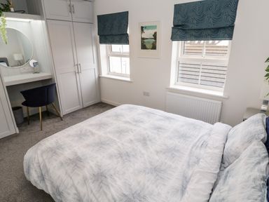 A bedroom with a bed, two windows with blue blinds, a wardrobe, and a desk with a mirror and chair at 20 Bishops Way in Falmouth