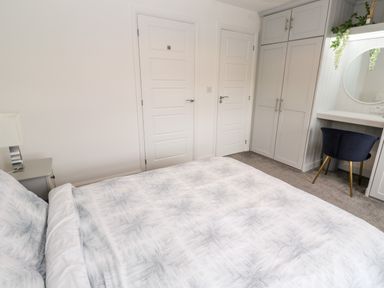 A bedroom with a bed covered in patterned bedding two white doors a bedside table with a lamp built in wardrobe and a dressing table with a round mirror and a dark chair at 20 Bishops Way in Falmouth