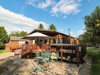 An outdoor patio area with a green metal table and chairs under a white umbrella beside a wooden lodge with a woodpile storage at Evergreen Lodge in Felmoor Holiday Park near Felton