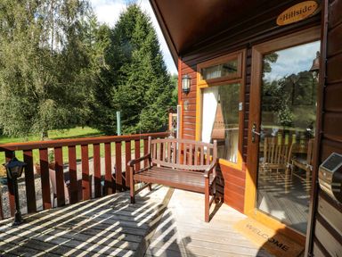 A wooden bench on a deck outside a cabin with glass door and railing at Evergreen Lodge in Felmoor Holiday Park near Felton