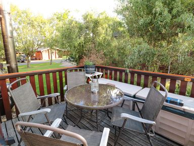 An outdoor wooden deck with a round glass table, four chairs, a bench with storage, and greenery around at Evergreen Lodge in Felmoor Holiday Park near Felton