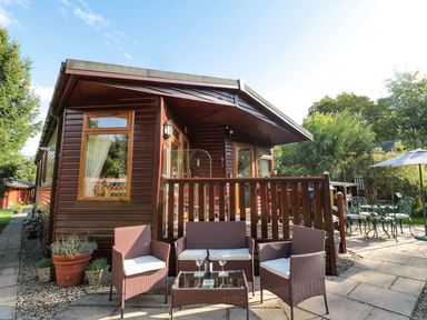 A wooden lodge with outdoor wicker chairs and a table with wine glasses and a bottle at Evergreen Lodge in Felmoor Holiday Park near Felton