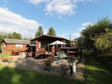 A wooden cabin with a patio table umbrella and chairs outside near trees at Evergreen Lodge Felmoor Holiday Park near Felton