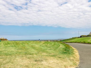 A grassy open area next to a paved pathway with the sea visible in the distance at Evergreen Lodge Felmoor Holiday Park near Felton