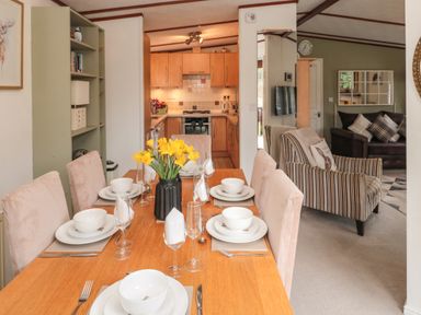 A dining table set with plates bowls glasses and a vase of yellow flowers with a kitchen in the background at Evergreen Lodge Felmoor Holiday Park near Felton