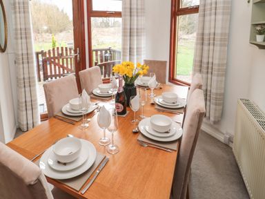 A dining table set for six with dishes glasses and a bottle of sparkling drink near large windows at Evergreen Lodge Felmoor Holiday Park near Felton