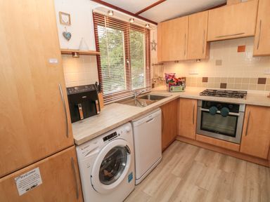 A kitchen with wooden cabinets a washing machine dishwasher oven gas stove and a window with blinds at Evergreen Lodge Felmoor Holiday Park near Felton