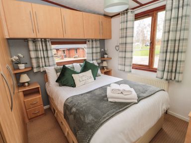 A bedroom with a bed set with green and white pillows and towels on top next to windows with plaid curtains at Evergreen Lodge Felmoor Holiday Park near Felton