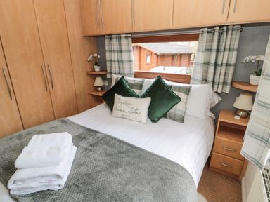 A bedroom with a double bed green and plaid cushions wooden cabinets bedside tables and folded towels on the bed at Evergreen Lodge Felmoor Holiday Park near Felton