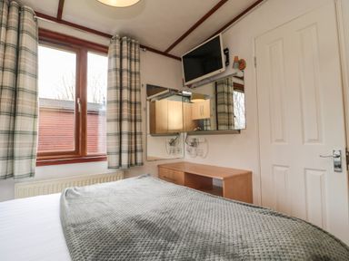 A bedroom with a bed a window with plaid curtains a wooden cabinet and a wall-mounted television at Evergreen Lodge Felmoor Holiday Park near Felton