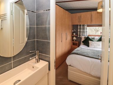 A bathroom sink with a round mirror and a bedroom with a bed green pillows and wooden wardrobes at Evergreen Lodge in Felmoor Holiday Park near Felton