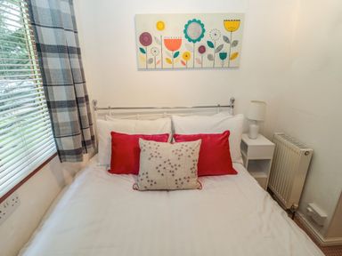 A bedroom with a double bed red and white cushions a bedside table with a lamp a radiator and a window with checkered curtains at Evergreen Lodge in Felmoor Holiday Park near Felton