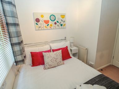 A bedroom with a bed white pillows red cushions bedside table with lamp and floral wall art at Evergreen Lodge Felmoor Holiday Park near Felton