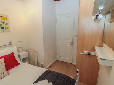 A bedroom with a bed red and beige cushions a white bedside table and lamp a door and a wooden wall panel with shelf and electrical outlets at Evergreen Lodge Felmoor Holiday Park near Felton