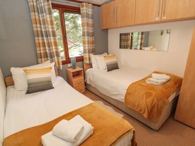 A bedroom with two single beds two pillows on each bed bedside table lamp wooden cupboards and window with curtains at Evergreen Lodge Felmoor Holiday Park near Felton