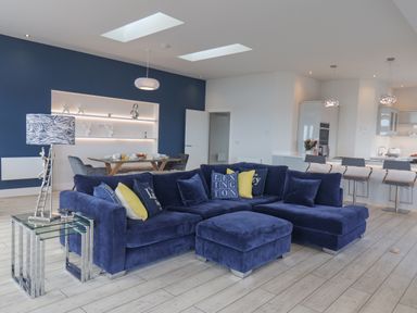 An open living area with a blue sectional sofa and cushions a dining table with chairs and a kitchen with bar stools at The Penthouse in Scarborough