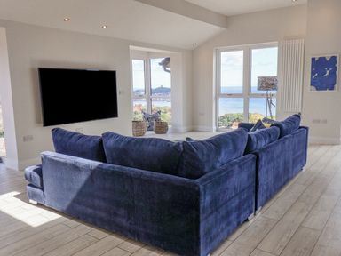 A living room with a blue sofa facing a wall-mounted TV and windows showing a sea view at The Penthouse in Scarborough