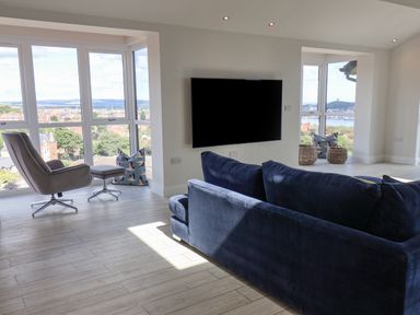 A living room with a blue sofa a wall mounted TV and a chair with an ottoman near large windows at The Penthouse in Scarborough