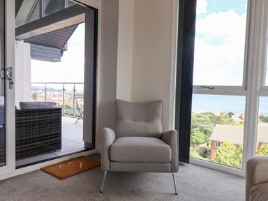 A grey armchair next to large windows overlooking houses and trees with a balcony visible outside at The Penthouse in Scarborough
