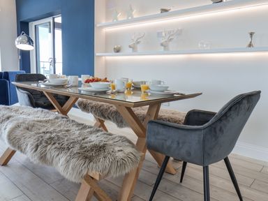A dining area with a glass top table set with dishes and orange juice with sheepskin covered benches and grey chairs at The Penthouse in Scarborough