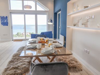 A dining area with a glass table set with dishes and drinks and sheepskin covered benches by large windows overlooking the sea at The Penthouse in Scarborough
