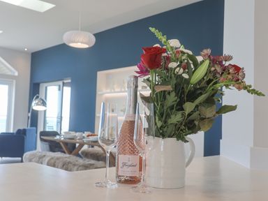 A kitchen counter with a bottle of rose wine two wine glasses and a vase of flowers with a dining table and blue walls in the background at The Penthouse in Scarborough