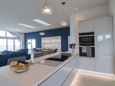 A modern kitchen with white cabinets and an island with cheese and grapes overlooking a living area and dining table at The Penthouse in Scarborough