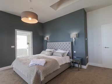 A bedroom with a large bed a gray padded headboard bedside tables and a door leading to a bathroom at The Penthouse in Scarborough