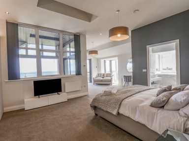 A bedroom with a bed pillows and folded towels a sofa a TV on a white stand and large windows overlooking the sea at The Penthouse in Scarborough