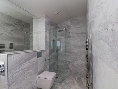 A bathroom with a wall-mounted toilet a glass shower enclosure and grey marble walls and floor at The Penthouse in Scarborough