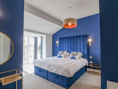 A bedroom with a blue upholstered bed and headboard two pillows side tables wall sconces and a round mirror at The Penthouse in Scarborough