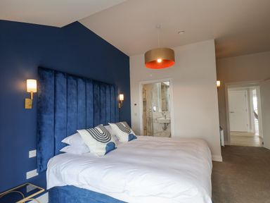 A bedroom with a large blue upholstered bed with white bedding and two pillows with patterns two wall lamps and a doorway to a bathroom at The Penthouse in Scarborough