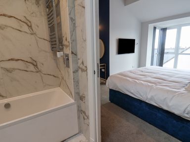 A bathroom with a bathtub and marble walls next to a bedroom with a bed a wall-mounted tv and a window at The Penthouse in Scarborough