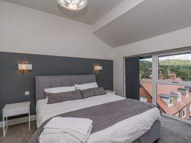 A bedroom with a double bed bedside tables wall lights and a window overlooking rooftops and trees at The Penthouse in Scarborough