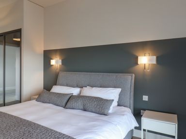 A bedroom with a gray upholstered bed white bedding two gray pillows two wall lamps and a white bedside table at The Penthouse in Scarborough