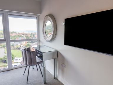 A room with a mirrored desk and grey chair next to a window with a view and a wall mounted television at The Penthouse in Scarborough