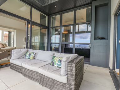 A wicker sofa with cushions on a balcony with glass windows and a black sculpture at The Penthouse in Scarborough