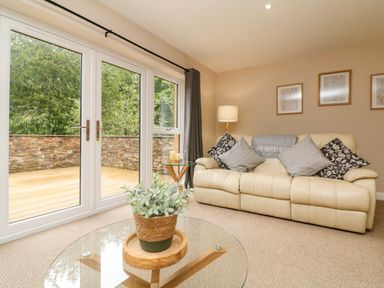 A living room with a cream sofa cushions a glass coffee table with a potted plant a side table with candles and a lamp near large windows at Spicery in Tedburn St Mary