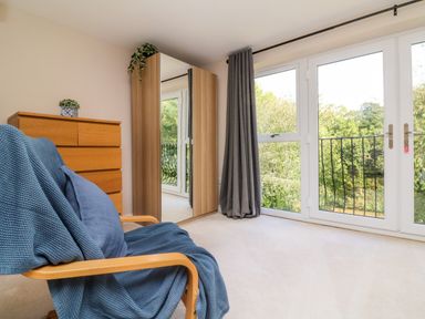 A room with a wooden chair covered with a blue blanket and pillow next to a wooden chest of drawers and a wardrobe with mirrored doors and large windows with grey curtains at Spicery in Tedburn St Mary