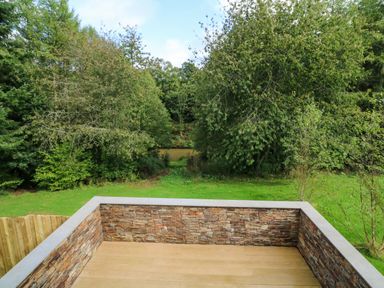 A wooden deck with stone walls overlooking a grassy yard and trees at Spicery in Tedburn St Mary