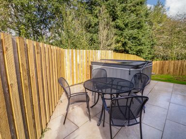 A patio with a black metal table and four chairs next to a hot tub and wooden fence at Spicery in Tedburn St Mary