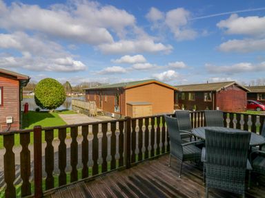 A wooden deck with a round table and four chairs overlooking wooden lodges and a small lake at Lakeside 20 in Warton near Carnforth
