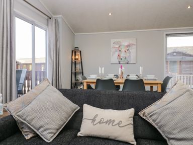 A living room with a dark sofa with cushions and a dining table set with plates glasses and a bottle at Lakeside 20 in Warton near Carnforth